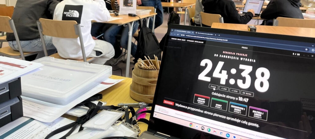 Laptop prowadzących z zegarem redakcji „Do zamknięcia wydania 24:38”, „Oddajecie strony o 10:47” i depeszą „Wydawca przypomina: strona pierwsza sprzedaje całą gazetę”; obok skrzynki z materiałami, w tle sala