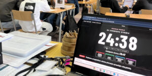 Laptop prowadzących z zegarem redakcji „Do zamknięcia wydania 24:38”, „Oddajecie strony o 10:47” i depeszą „Wydawca przypomina: strona pierwsza sprzedaje całą gazetę”; obok skrzynki z materiałami, w tle sala