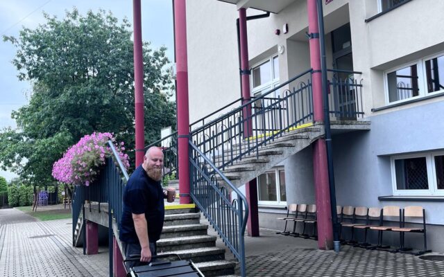 Prowadzący z walizką Przenośnej Redakcji przed wejściem do Szkoły Podstawowej w Gorzkowie