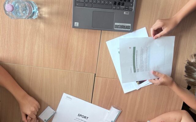 Widok z góry na stolik działu Sport: karta działu, identyfikatory, laptop i ręce uczniów nad materiałami