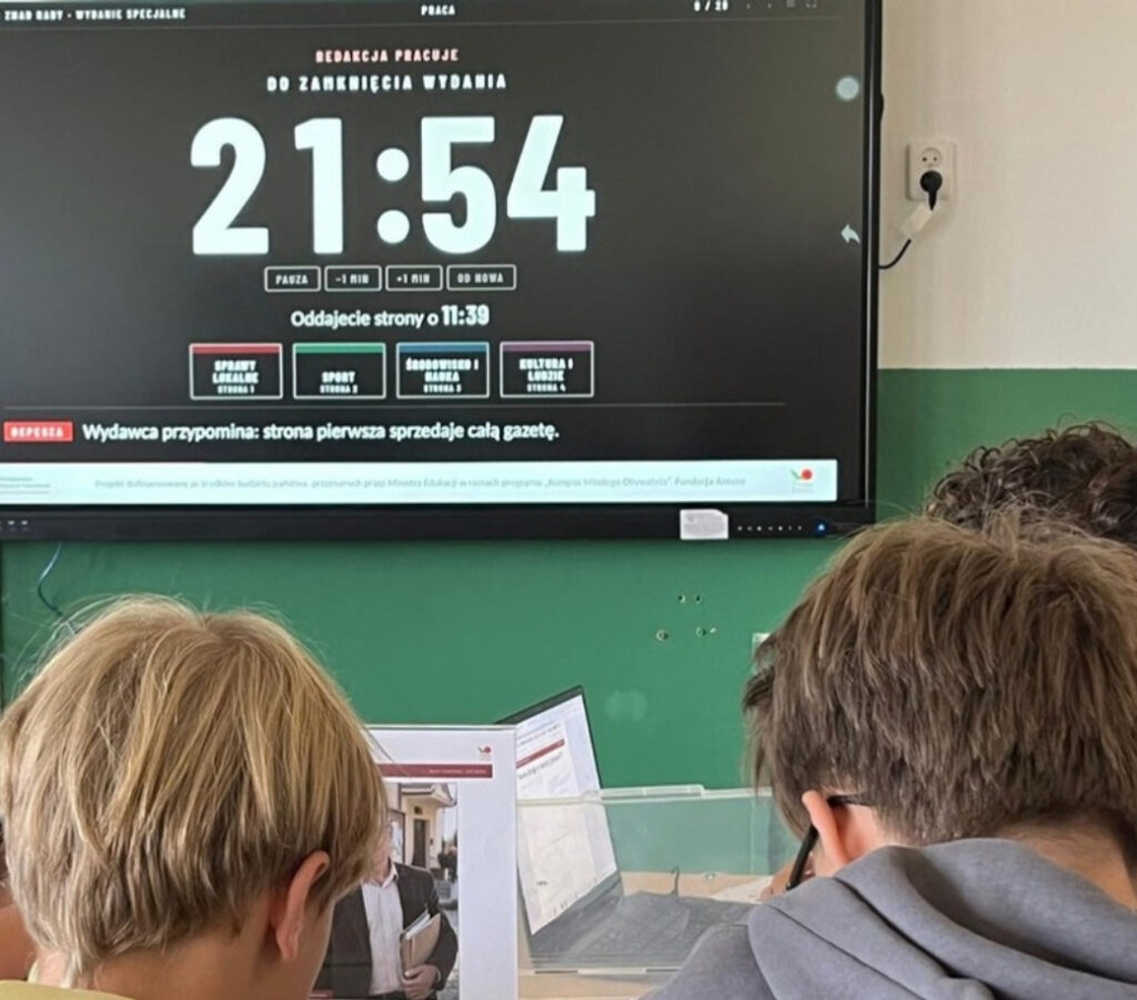 Ekran w sali z zegarem redakcji „Do zamknięcia wydania 21:54” i nazwami czterech działów, przed nim uczniowie widziani od tyłu