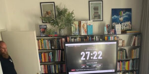Czytelnia Biblioteki Pedagogicznej w Bochni: między regałami z książkami ekran z zegarem redakcji „Do zamknięcia wydania 27:22”, w drzwiach prowadzący