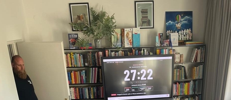 Czytelnia Biblioteki Pedagogicznej w Bochni: między regałami z książkami ekran z zegarem redakcji „Do zamknięcia wydania 27:22”, w drzwiach prowadzący