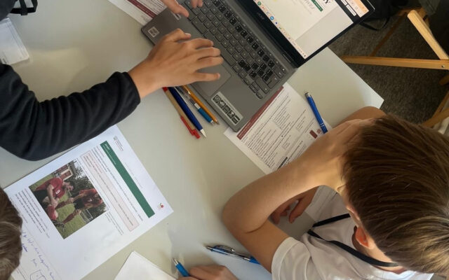 Widok z góry na stolik działu Sport: laptop z szablonem „Kuriera znad Raby”, karta szefa działu, materiały i ręce uczniów