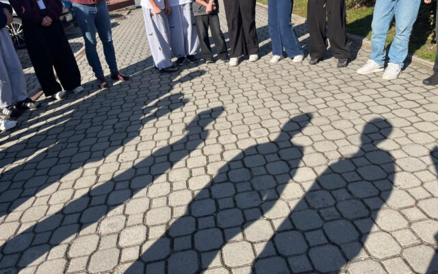 Cienie stażystów na kostce brukowej przed biblioteką, uczniowie widoczni od pasa w dół